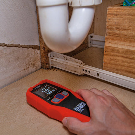 Action shot showing a hand placing the ET140 moisture meter on the base of a wooden cabinet under a sink. The meter displays a relative moisture content of 7.8 percent with green LED bars illuminated on the graph.