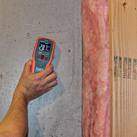 Action shot of a person using the ET140 moisture meter to test a concrete basement wall next to exposed pink fiberglass insulation and a wooden wall stud. The device is set to masonry mode and displays a 28.7 percent relative moisture reading with yellow LED bars illuminated.