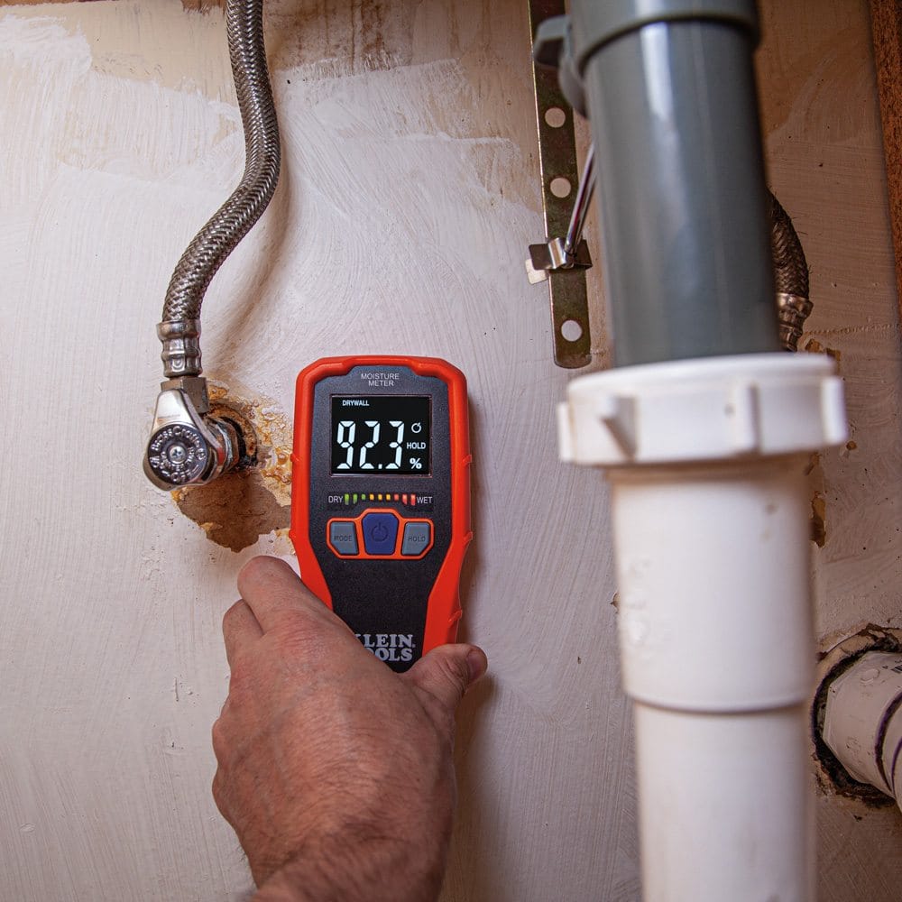 Action shot of a worker using the ET140 pinless meter to check for leaks under a sink. The device is held against a white wall near plumbing pipes and shows a high relative moisture reading of 92.3 percent with the red LED bars illuminated.