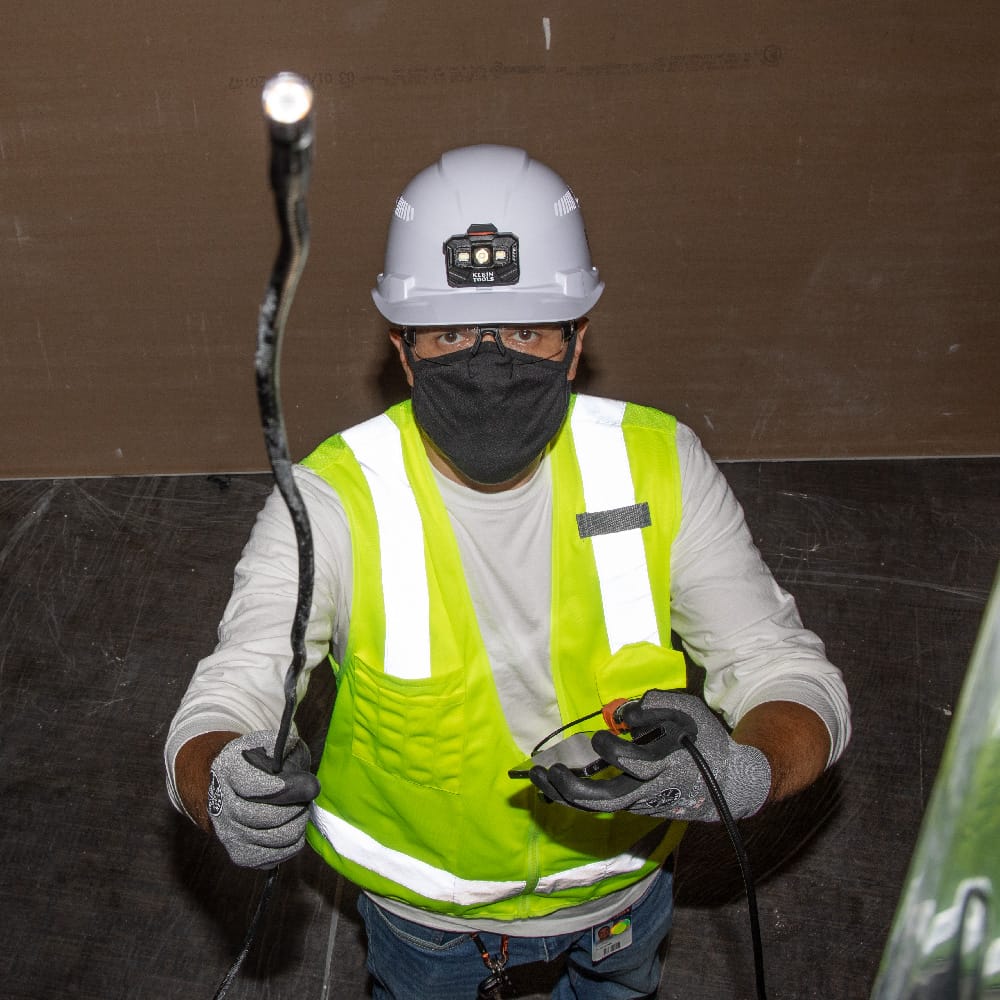 A professional technician wearing a hard hat, safety glasses, and high-visibility vest using the ET16 borescope. The technician holds the armored gooseneck probe to inspect overhead or inaccessible areas while monitoring the live video feed on an Android smartphone.