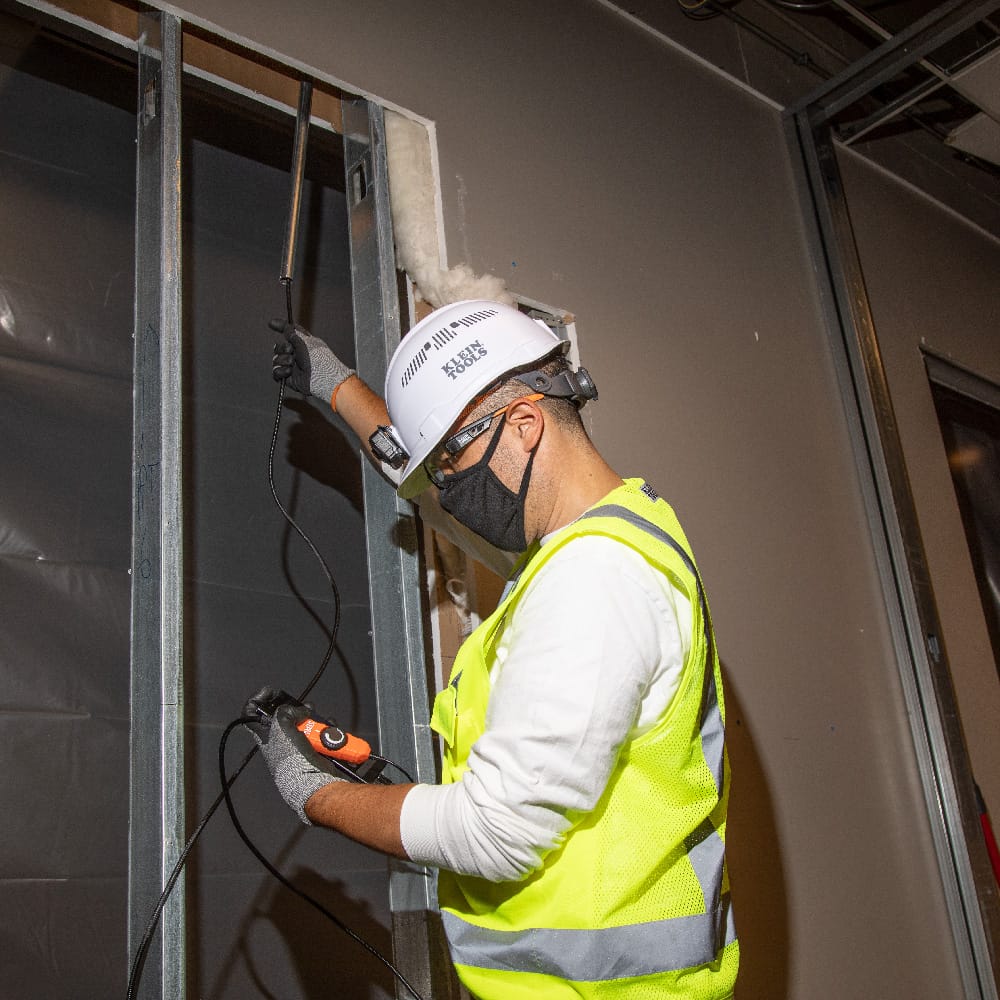 Action shot of an electrician using the ET16 inspection camera to look inside a wall cavity during a commercial construction project. The 10-foot gooseneck is shown extended through a metal stud frame, demonstrating the tool's shape retention and reach.