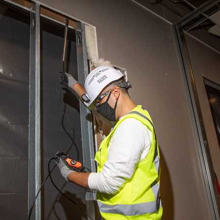 Action shot of an electrician using the ET16 inspection camera to look inside a wall cavity during a commercial construction project. The 10-foot gooseneck is shown extended through a metal stud frame, demonstrating the tool's shape retention and reach.