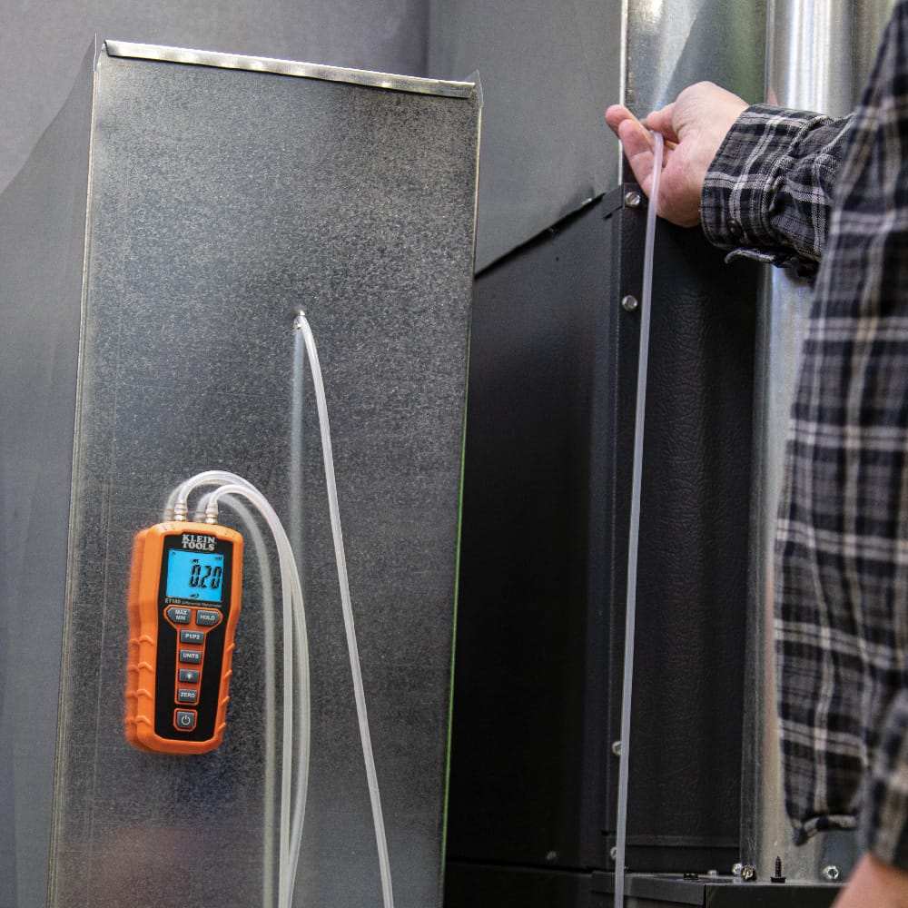 Action shot demonstrating the hands-free capability of the Klein Tools ET180. The digital manometer is secured to a galvanized steel air duct via its internal magnet, allowing the technician to use both hands to manipulate the pressure probe during a static pressure test.