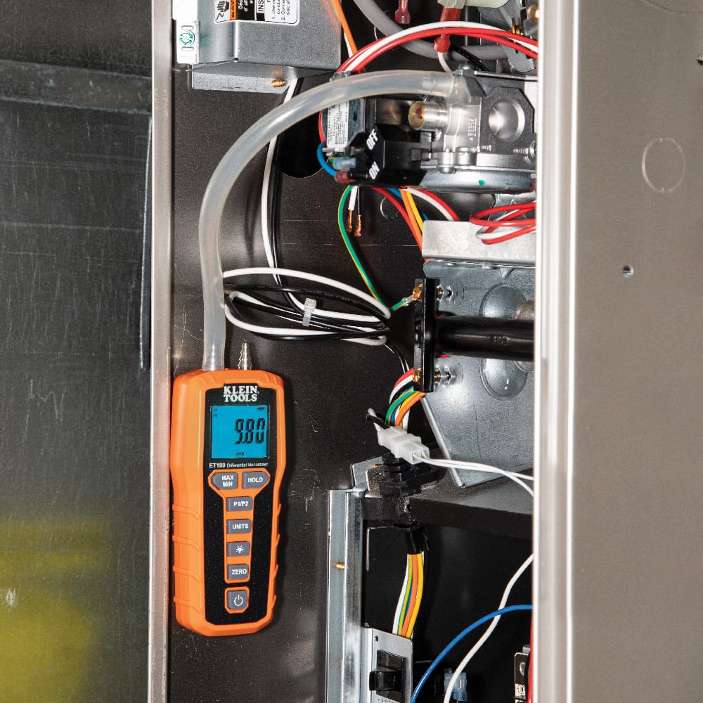Close-up of the Klein Tools ET180 manometer being used to troubleshoot an HVAC furnace system. The meter is secured to the metal furnace cabinet using its integrated rear magnet, providing hands-free operation while measuring gas manifold pressure through a silicon tube connection, showing a clear reading of 9.80 on the backlit blue display.