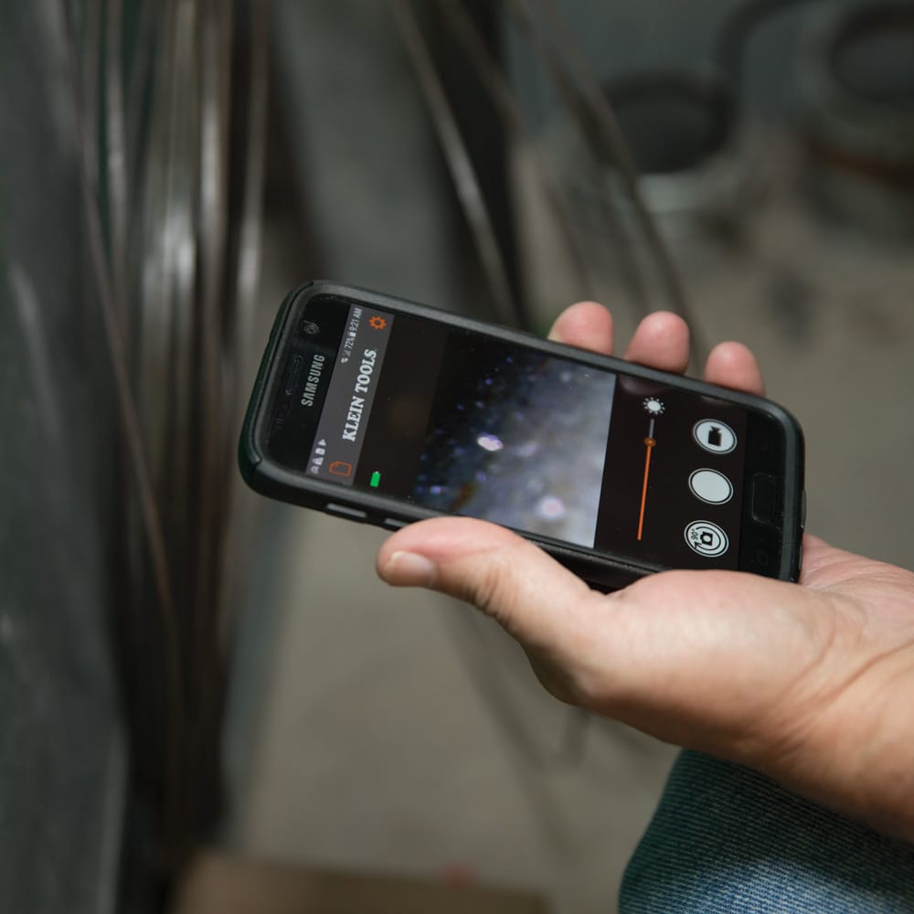 Close-up action shot of a technician holding a smartphone running the Klein Tools Borescope App. The screen displays a live, high-clarity view of internal wiring being inspected with the ET20 WiFi borescope, highlighting the ease of capturing and sharing diagnostic images.