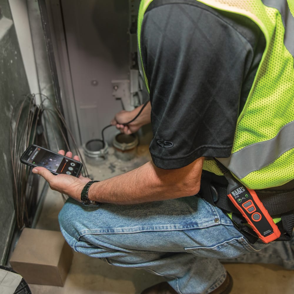 Action shot of a professional technician using the ET20 WiFi borescope to inspect a floor drain. The technician monitors a real-time video feed on an Android smartphone while the transmitter is clipped to their belt, demonstrating the wireless freedom and professional utility of the tool.