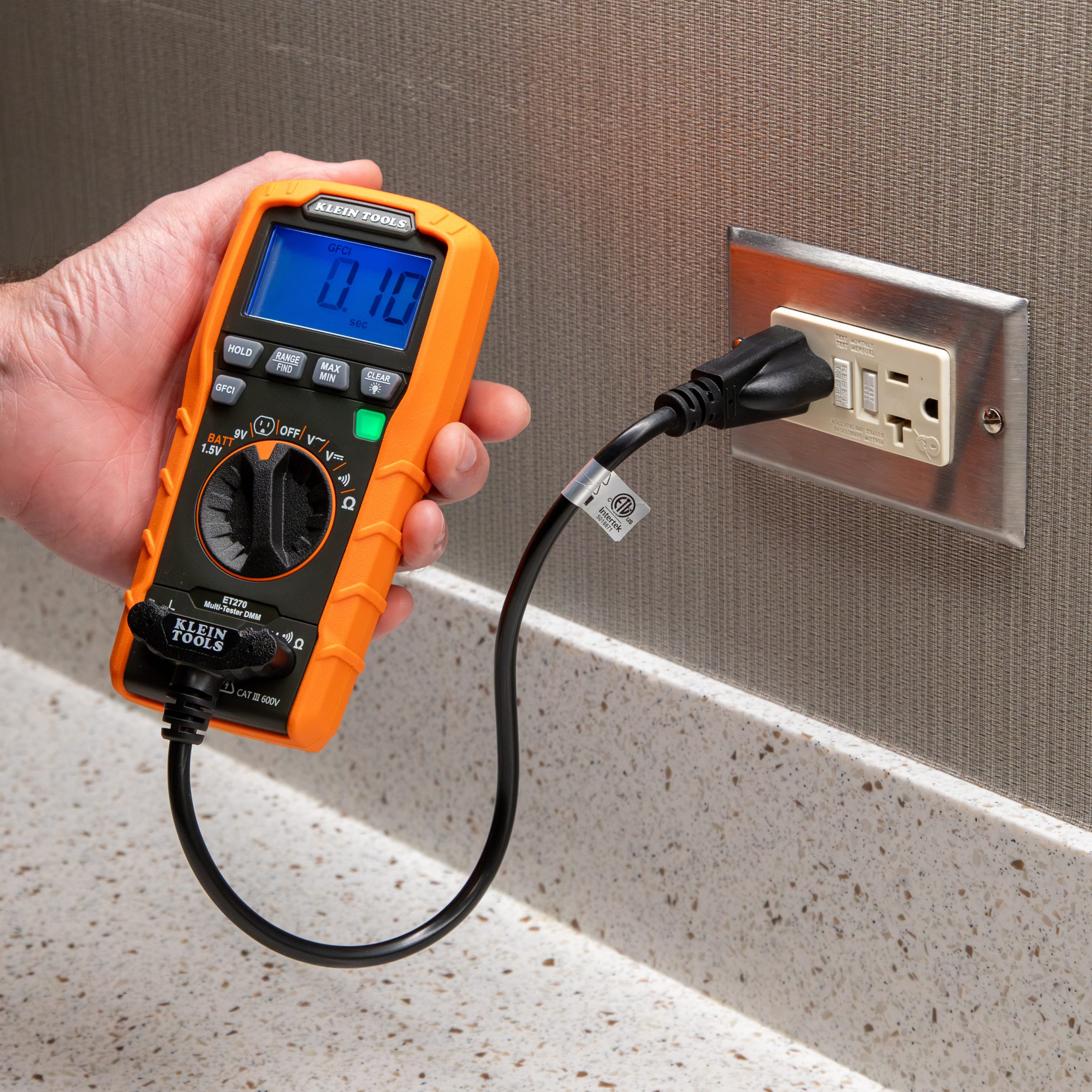 Close-up of the Klein Tools ET270 performing a GFCI trip test on a kitchen or bathroom style receptacle. The user is pressing the dedicated GFCI button, and the high-contrast display shows a trip time of 0.10 seconds, confirming the safety device is operating within specifications.