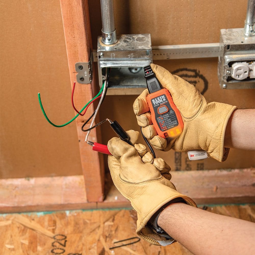 A person wearing leather gloves holding the transmitter connected to the alligator clip adapter, which is attached to bare wires inside an open wall junction box.