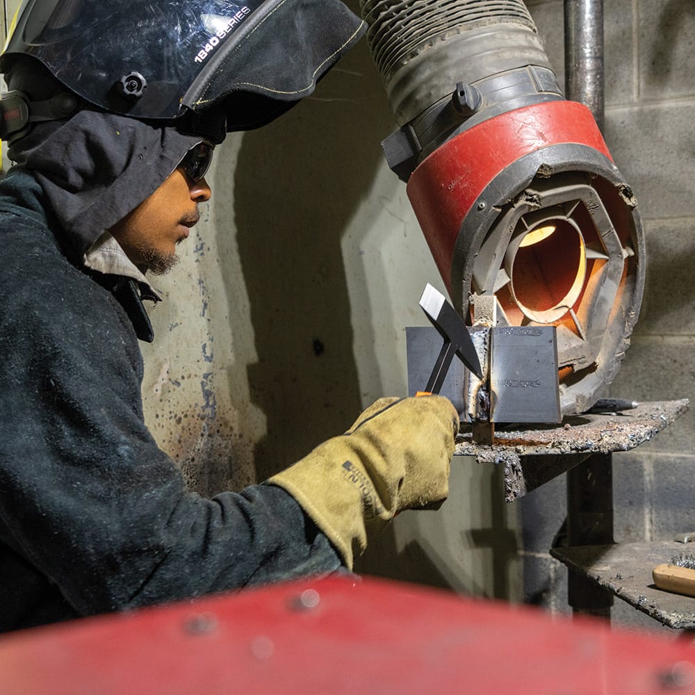 Welder using protective gear in a workshop with a Klein Tools H80612 Welder’s Chipping Hammer nearby, emphasizing slag removal tools used during metal fabrication and post‑weld cleanup.
