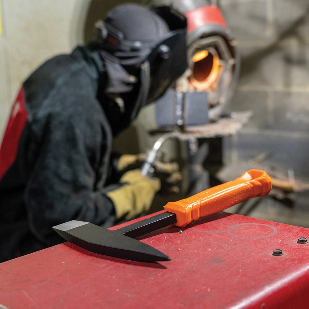 Welding scene with a Klein Tools H80612 Welder’s Chipping Hammer resting on a red surface in the foreground while a welder works with protective gear and high‑heat equipment.