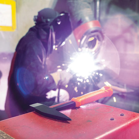 Active welding environment with sparks flying and a Klein Tools H80612 Welder’s Chipping Hammer on a workbench, used for slag removal and post‑weld cleanup in metalworking.