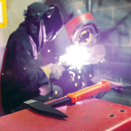 Industrial welding scene with bright sparks and a Klein Tools H80612 Welder’s Chipping Hammer positioned on a red work surface, highlighting essential slag removal tools for welders.