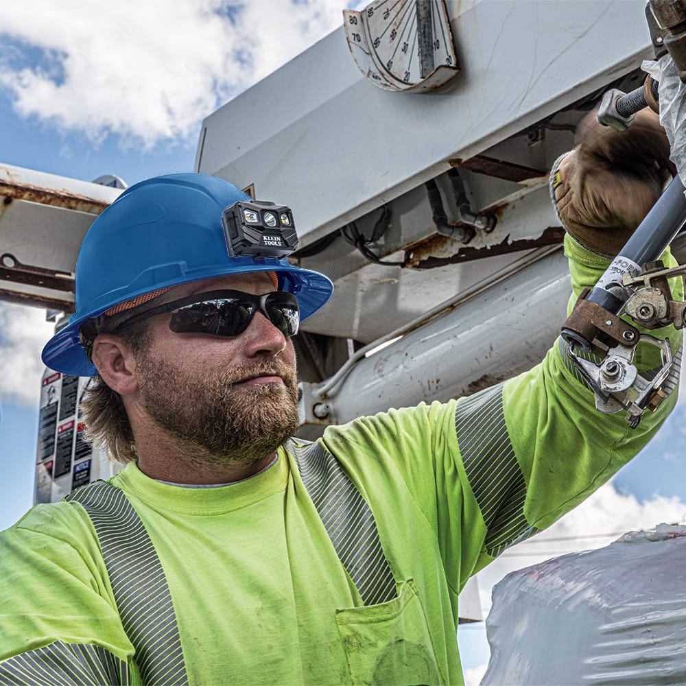 Utility technician wearing a Klein 60249 blue full brim hard hat with a front-mounted LED headlamp while servicing overhead electrical equipment.