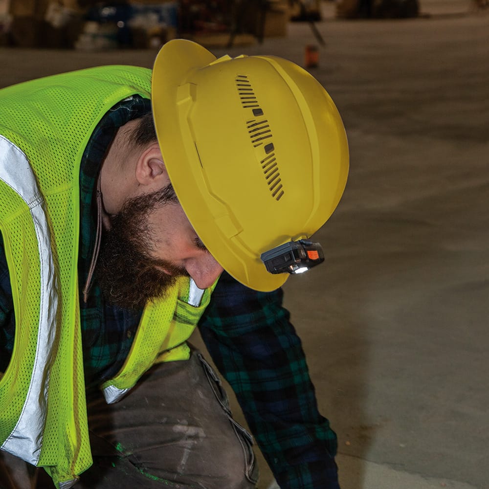 Contractor wearing a Klein 60262 yellow vented hard hat with a front-mounted LED headlamp inspecting low-light commercial floor surfaces.