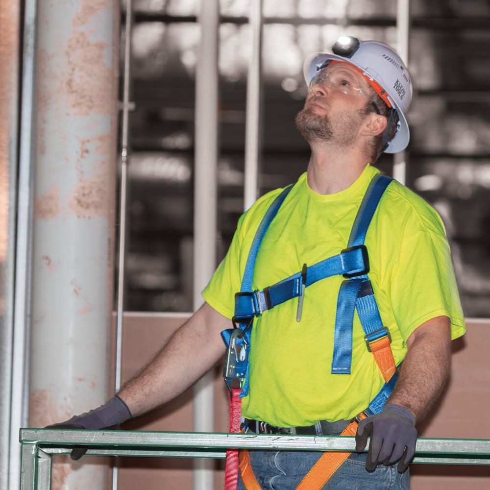 Construction tradesman wearing a Klein 60401 full brim hard hat equipped with a front-mounted LED headlamp while working in a safety harness on scaffolding.