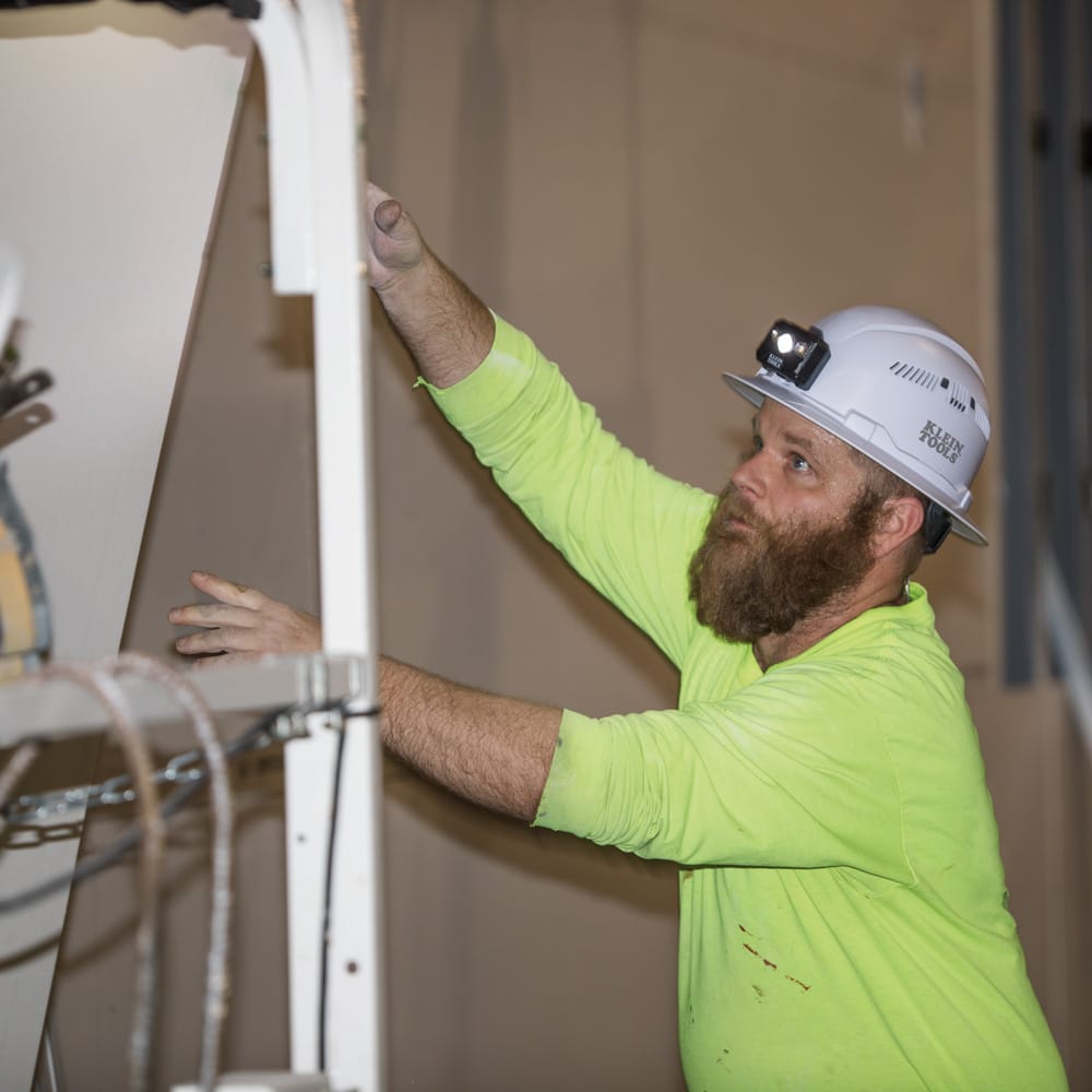 Worker wearing a Klein 60401 white vented hard hat with an attached headlamp positioning drywall panels in a commercial building interior.