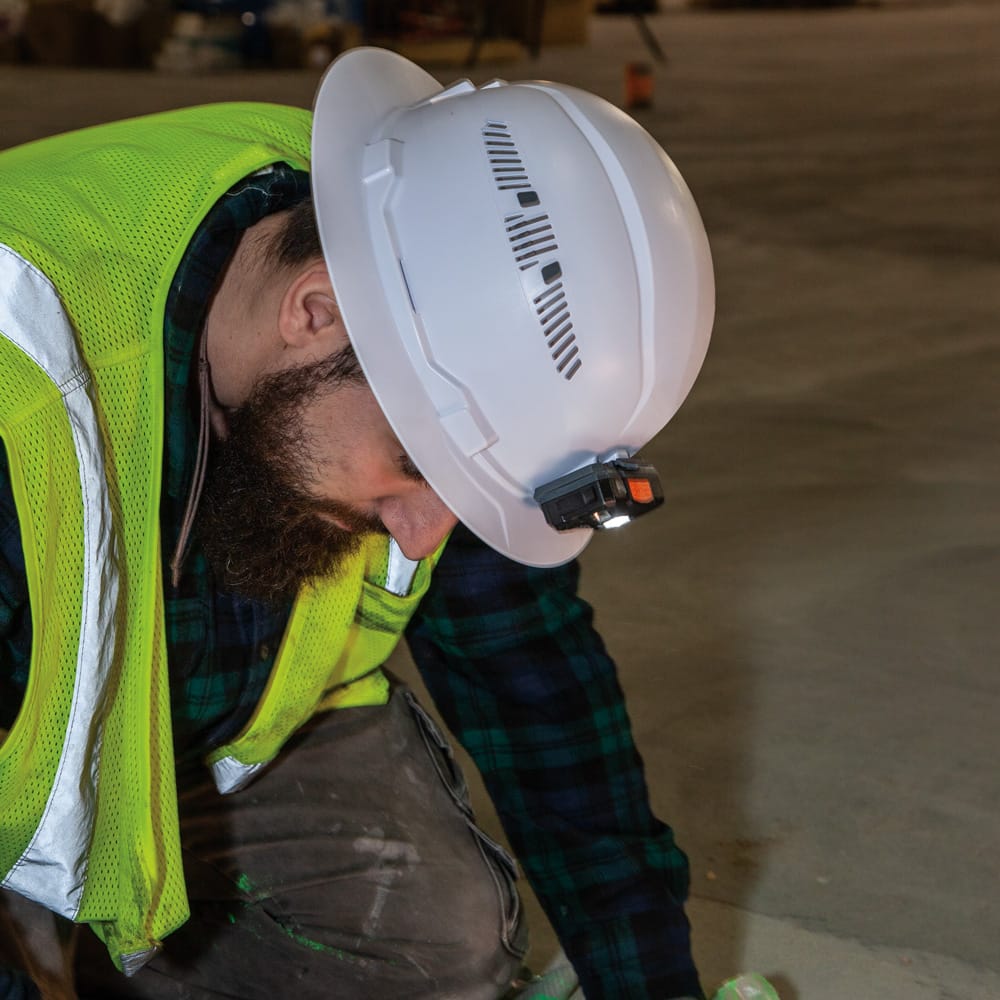 Contractor kneeling on a job site wearing a Klein 60401 full brim hard hat with a front-mounted headlamp providing hands-free surface illumination.