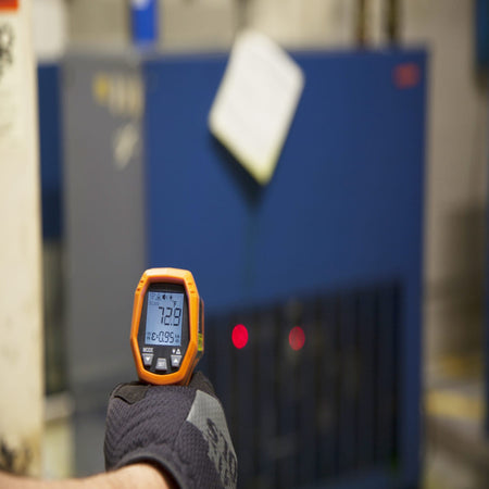 Action shot of the Klein Tools IR5 being used by a technician to measure the temperature of industrial machinery. The dual targeting lasers are visible as red dots on the blue equipment, and the meter display shows a real-time scanning measurement of 72.8 degrees Fahrenheit.