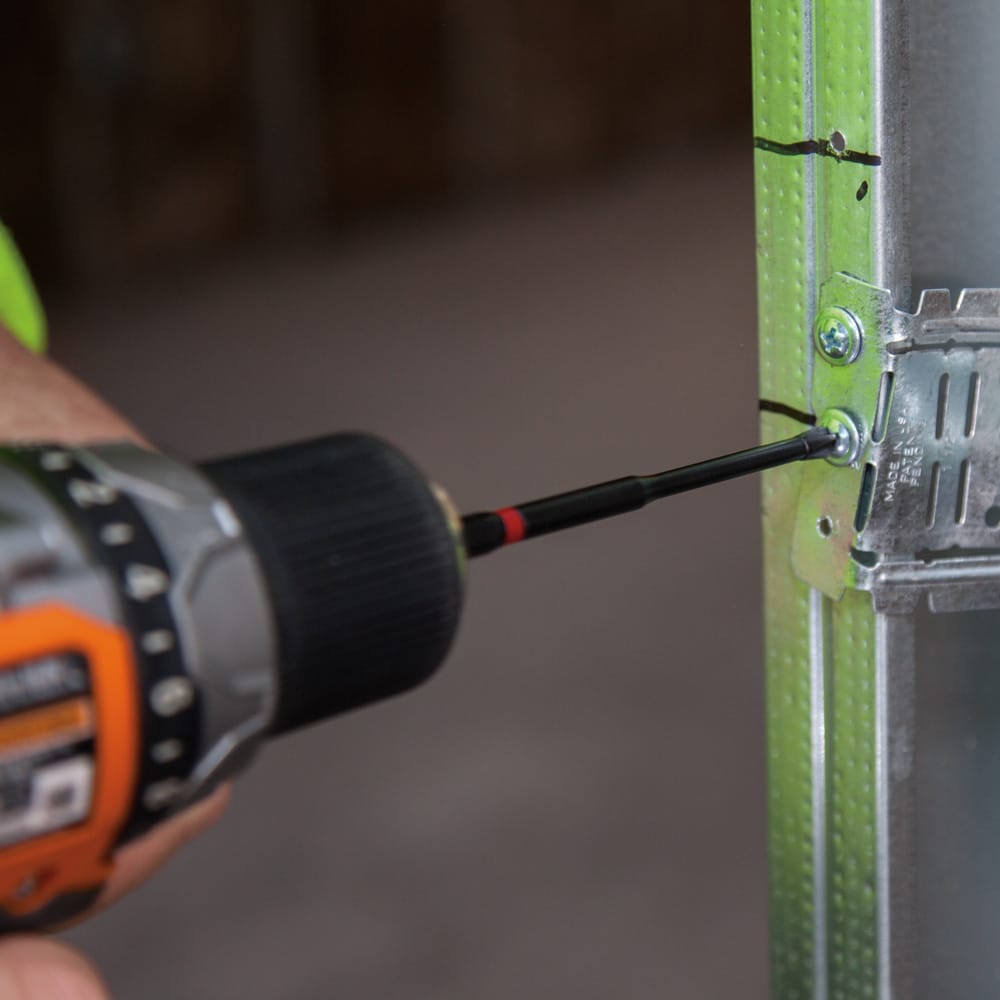 Technician using a power drill with a Klein 4-inch power bit to drive a mounting screw into a metal stud conduit bracket.