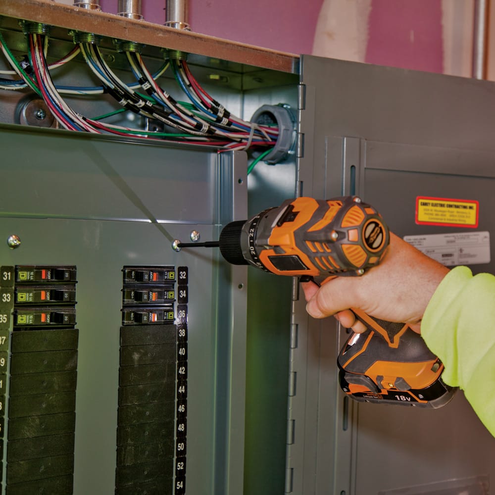 Electrician using a cordless impact driver fitted with a 4-inch Klein power bit to fasten a deadfront screw on an electrical panel.