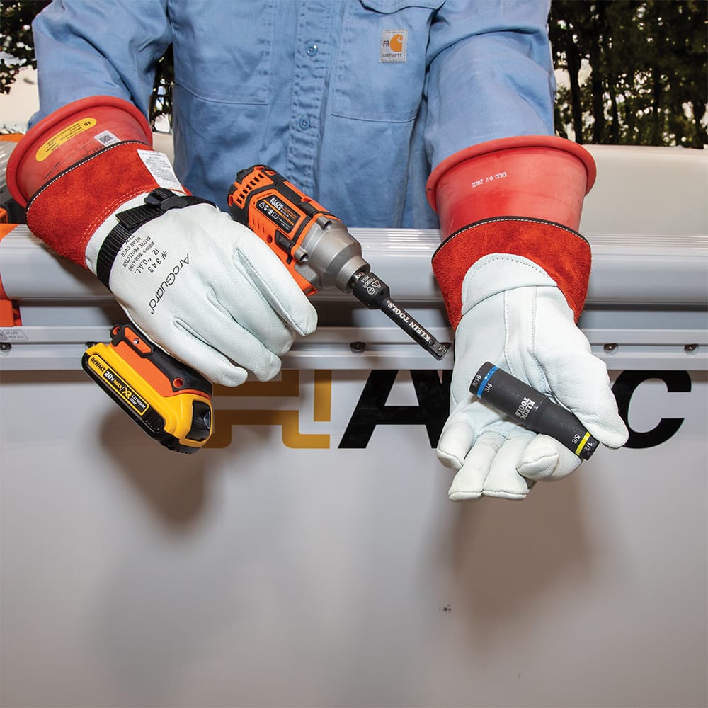 Lineman in high-voltage safety gloves holding a cordless impact driver fitted with a Klein 1/2 to 3/8-inch adapter beside a quad socket.