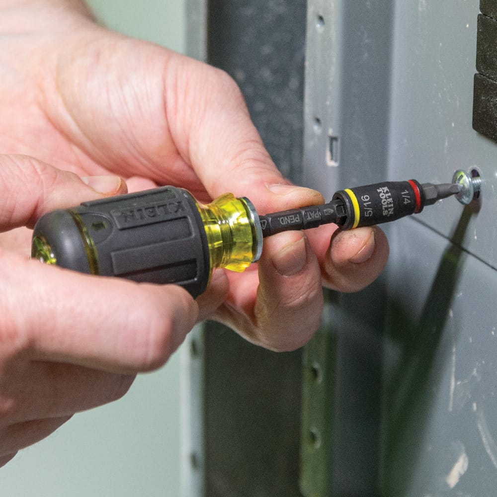 Technician using a manual Klein stubby driver fitted with the 32766 flip socket and a Phillips bit to adjust electrical enclosure cover screws.