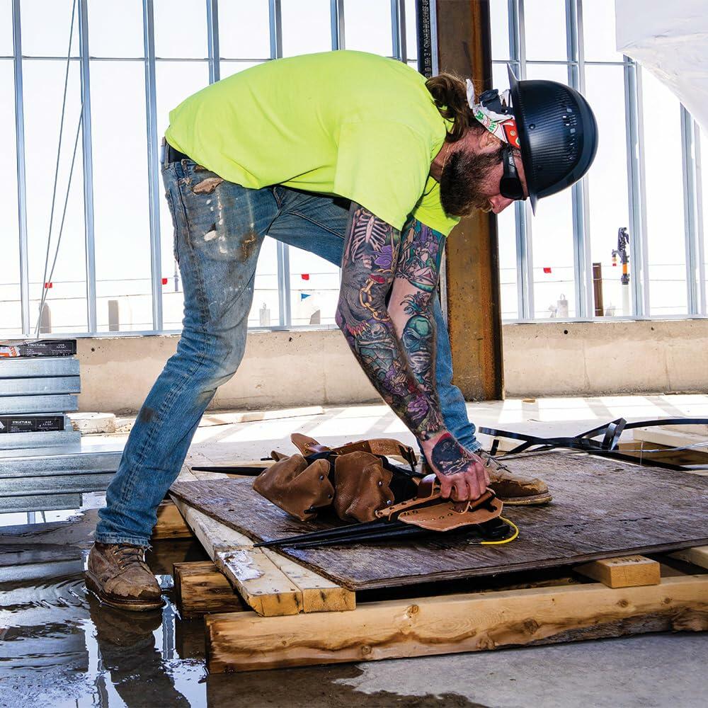 A construction worker wearing the Klein Tools Ironworker Tool Belt kneels to organize tools on a piece of plywood on a wet floor inside a building frame.