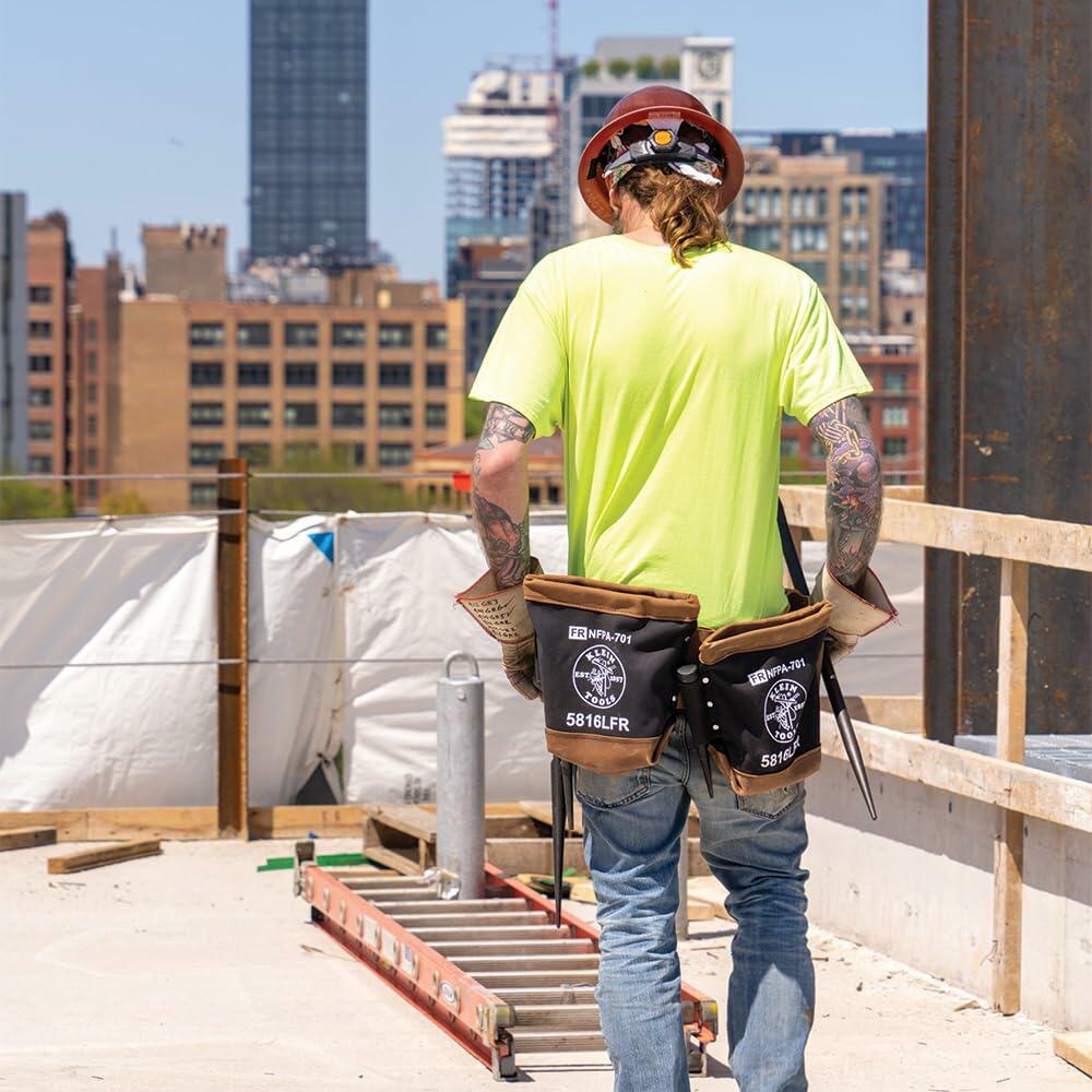 Rear view of a worker on a rooftop construction site wearing the Klein Tools Ironworker Belt System with two large flame-resistant bolt bags (5816LFR) hanging from the belt.
