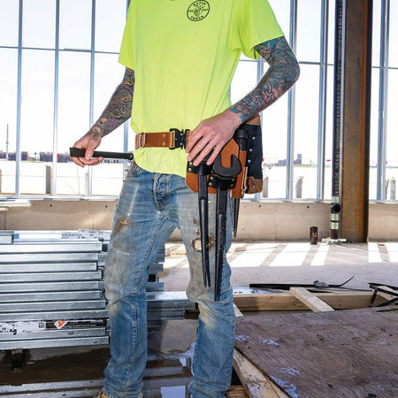 A worker wearing the Klein Tools Tool Belt reaches for a spud wrench or connecting bar stored securely in a leather holder on the belt, surrounded by construction materials.