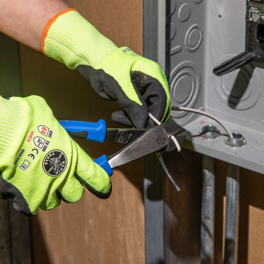 A detailed action view of a tradesman utilizing the Klein J200048 heavy-duty pliers to trim wire near a conduit fitting. The photo highlights the mechanical advantage provided by the high-leverage design and the comfort of the Journeyman handles during repetitive electrical work.