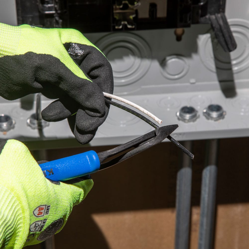 A detailed action shot of a professional technician wearing high-visibility safety gloves, using the Klein Tools J200048 8-inch heavy-duty diagonal cutting pliers. The image demonstrates the high-leverage angled head snipping electrical wire at a residential service panel, highlighting the tool's precision in tight spaces.