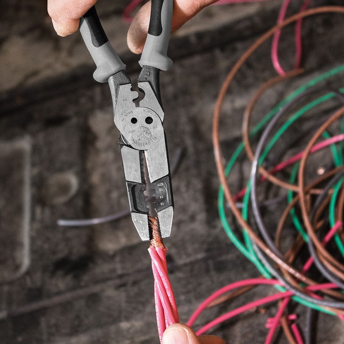 A close-up jobsite action view of an operator using the crimper die nest of the high-leverage hybrid pliers to securely compress and terminate a pink insulated wire conductor terminal connector over a pile of loose multi-colored electrical wiring paths.