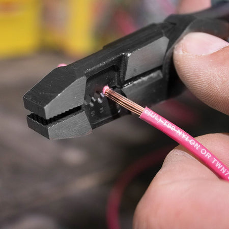A macro action shot focusing on the precision-ground wire stripping nests of the hybrid combo tool as it scores and strips away the pink outer nylon jacket insulation from a solid copper electrical wire thread, highlighting burr-free conductor wire preparation.