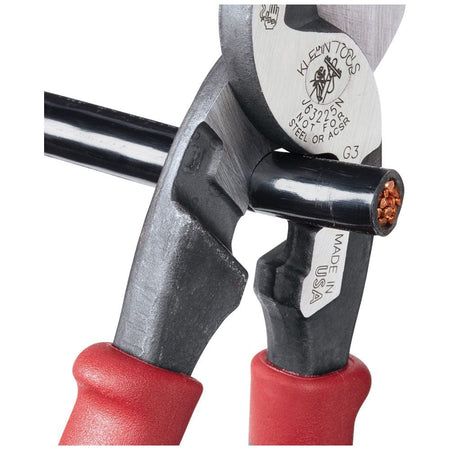 An action photograph of the Klein J63225N cable cutter performing a precise one-handed shear cut through a thick, black-insulated copper power cable.