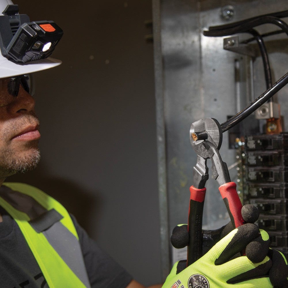 A tradesperson wearing a hard hat with a mounted LED light and high-visibility gear using the Klein J63225N high-leverage cutter to manage thick black cabling inside a commercial electrical panel.