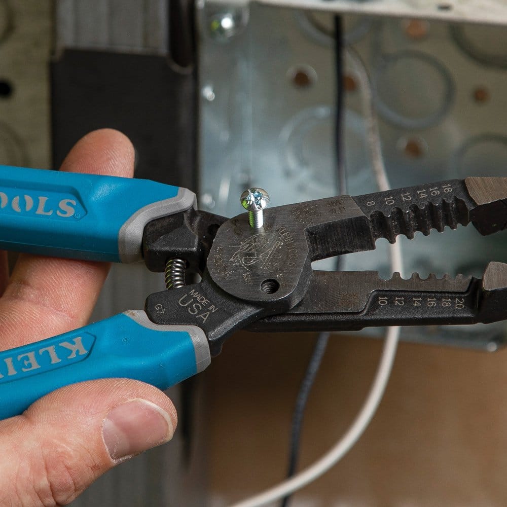 A detailed close-up of a machine screw being threaded into the integrated shearing hole of the Klein K12035 forged wire stripper, demonstrating the tool's ability to prep hardware for electrical boxes.
