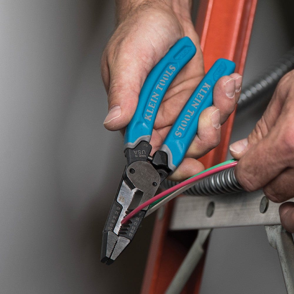 An action shot showing an electrician on a ladder using the Klein K12035 multi-tool to strip wires protruding from a metal conduit, highlighting its effectiveness in real-world jobsite environments.