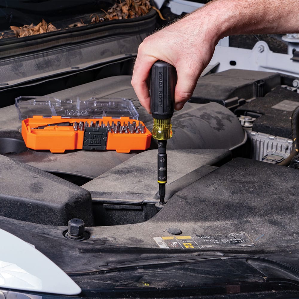 Mechanic using the Klein 32501HD 5/16-inch nut driver attached to a Cushion-Grip handle to turn automotive panel fasteners under an open hood.