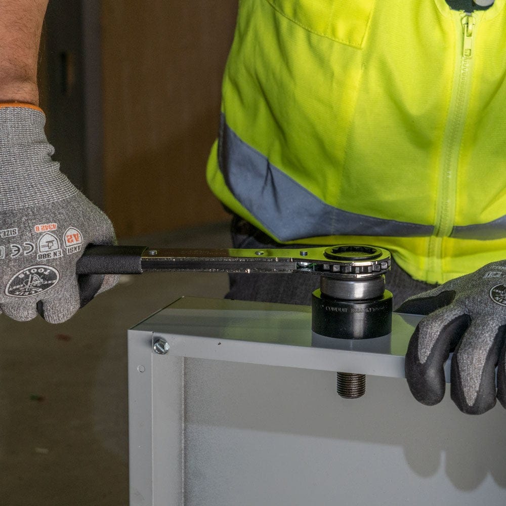 A worker in gloves and a safety vest uses the Klein ratcheting wrench to tighten a knockout punch on a metal electrical box.