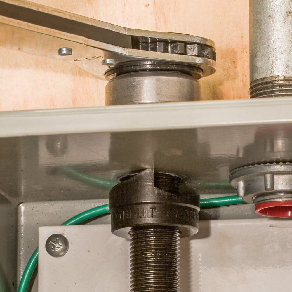 A close-up of the Klein knockout punch cutting through the wall of a metal enclosure, with the ratcheting wrench on top.