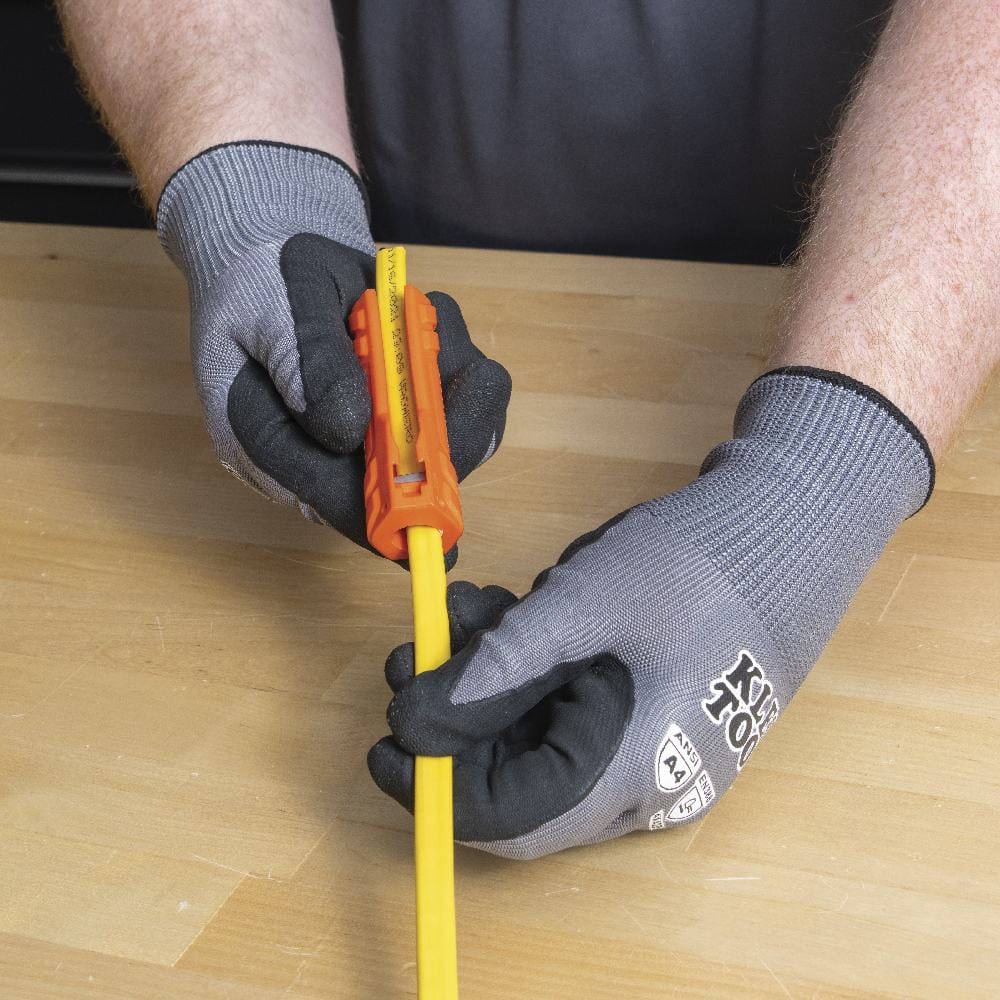 Gloved hands holding the orange multi-tool and inserting a yellow 12/2 NM Romex cable into the ripping slot to prepare for jacket removal.