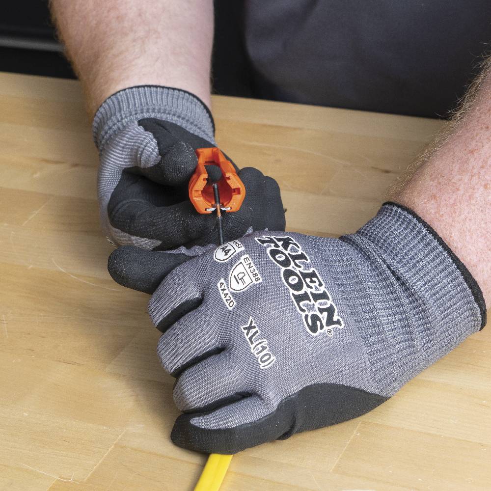 Gloved hands using the wire stripper notch on the end of the orange tool to remove insulation from a black 12 AWG conductor wire.