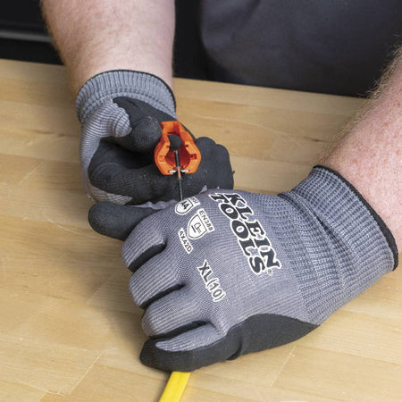 Gloved hands using the wire stripper notch on the end of the orange tool to remove insulation from a black 12 AWG conductor wire.
