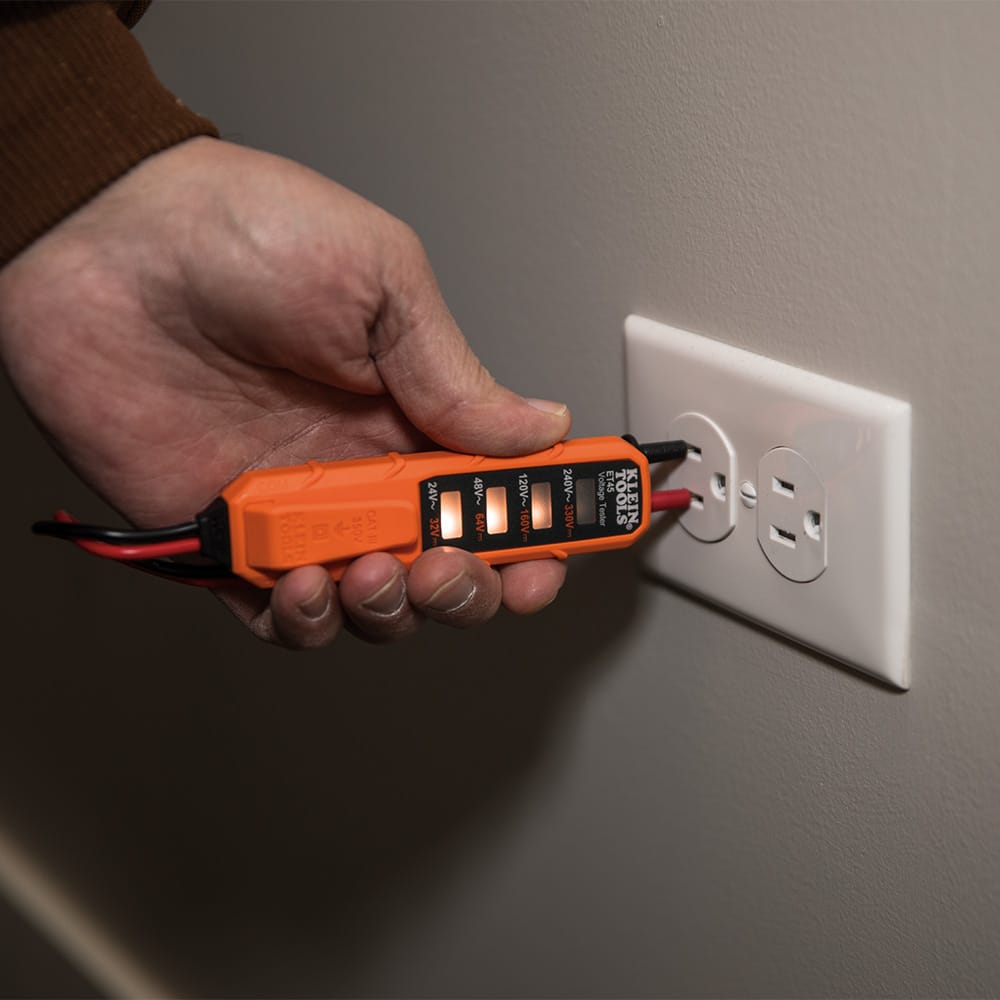 A professional action shot of a technician using the Klein Tools MM320 digital multimeter to verify voltage at a standard residential electrical outlet. The multimeter's high-contrast LCD displays the reading while the technician, wearing protective gear, uses the precision test leads to ensure circuit safety.