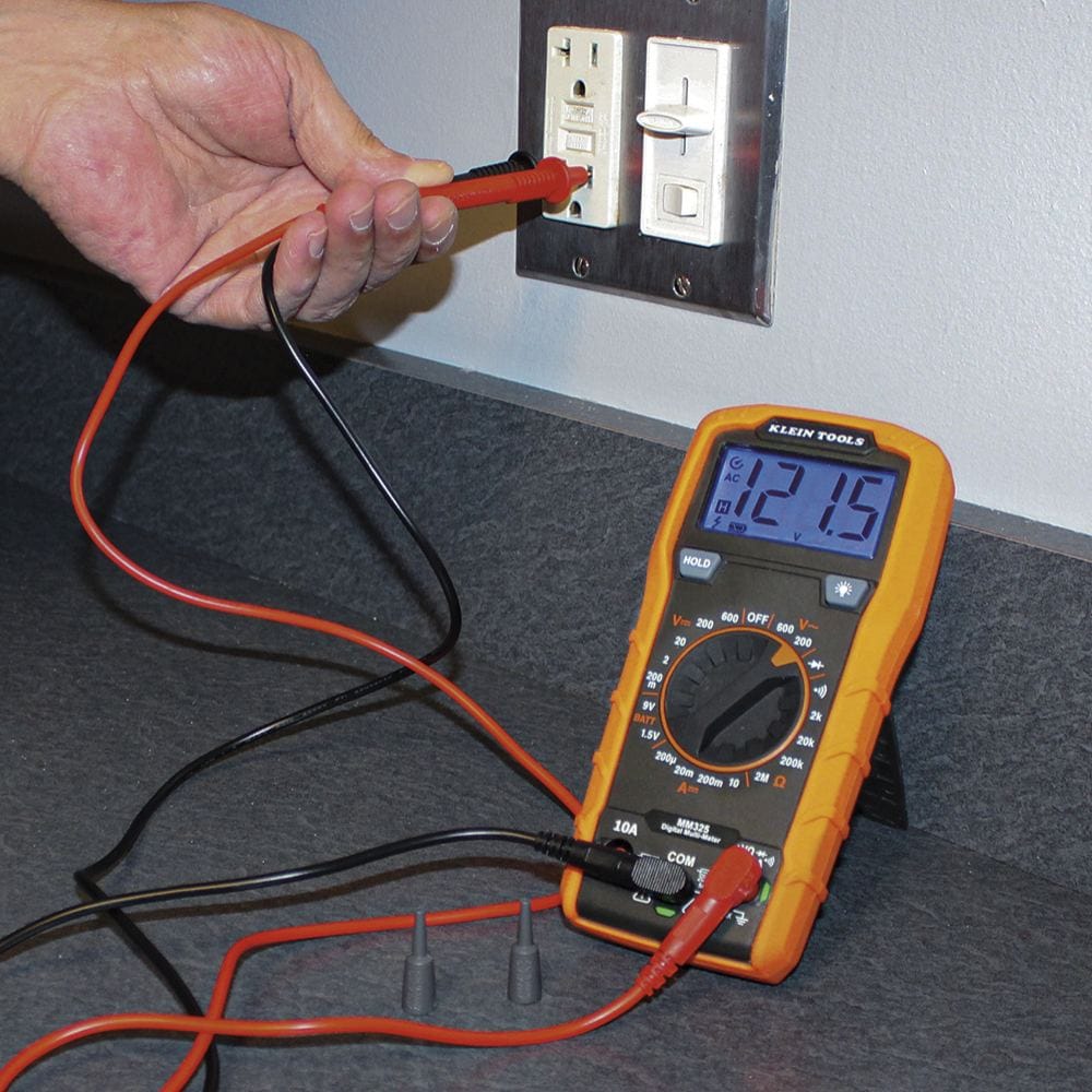 Action shot of a person using the MM325 multimeter to test a wall outlet. The multimeter is propped up on its kickstand on a grey countertop, showing a reading of 121.5V on its backlit blue display as the user inserts the red and black probes into an electrical socket.