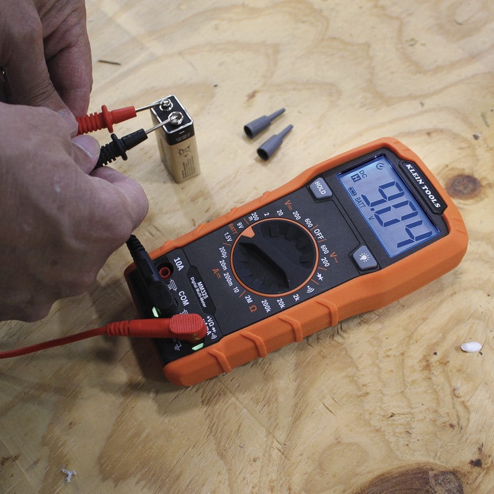 A person's hands are shown using the red and black probes of the Klein Tools MM325 digital multimeter to test a small 9V battery on a wooden surface. The multimeter's backlit blue screen displays a reading of 9.04.