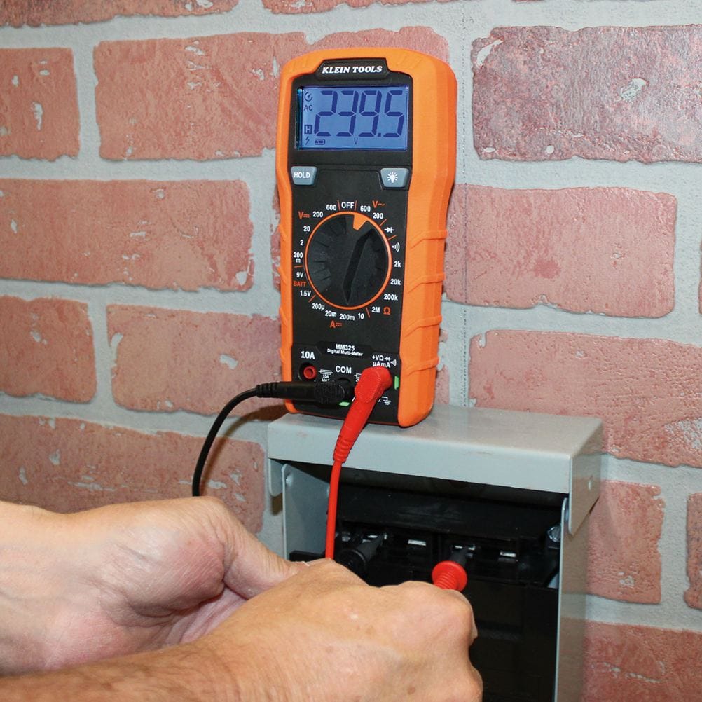 A worker uses the red and black probes of the MM325 multimeter to test a circuit inside an open grey electrical box mounted on a brick wall. The multimeter is propped up on its kickstand on top of the box, showing a reading of 239.5 on its backlit blue display.