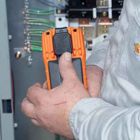 A close-up view of a worker's hands holding the back of the orange Klein Tools MM325 multimeter, demonstrating the installation or use of an optional black magnetic hanger accessory. The background shows blurred electrical wiring and components within a panel.