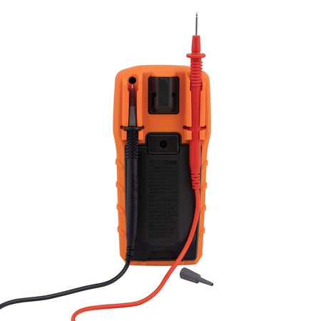Rear view of the MM325 multimeter showing the back of the orange and black housing. One red test lead is clipped into a dedicated storage slot on the right side, and a grey safety cap for a test lead probe lies on the white surface below.