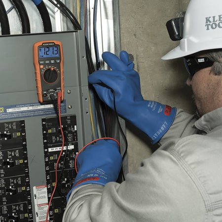 An electrician wearing protective rubber insulated gloves, a hard hat, and safety glasses uses the Klein Tools MM420 TRMS Multimeter to test voltage in a residential breaker panel. The multimeter is shown measuring $121.3V$ AC, demonstrating its practical use in live electrical troubleshooting and maintenance.